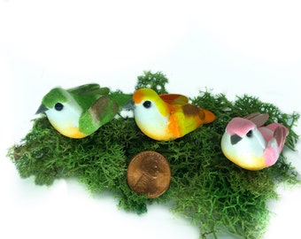 May include: Three small decorative birds made of felt, sitting on a bed of green moss. The birds are green, yellow, and pink with white bellies and black eyes. A copper penny is in the foreground.