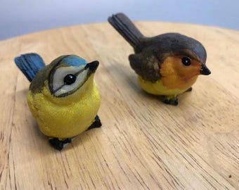 May include: Two decorative bird figurines. One is yellow with a blue head and tail. The other is brown, orange, and yellow. Both have black beaks and feet. They are placed on a wooden surface.
