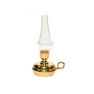 May include: A miniature oil lamp with a clear glass chimney and a brass base. The lamp features a rounded base, a small handle, and a gold finish. The glass chimney is elongated and tapers towards the top.