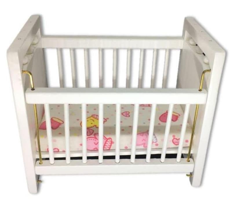 Puede incluir: Cuna de madera blanca con un colch&oacute;n estampado en blanco y rosa. La cuna tiene barras de metal dorado en los lados.