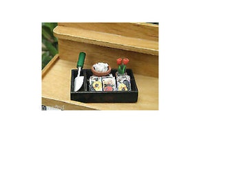 Könnte beinhalten: Ein Miniatur-Gartensatz in einer schwarzen Schale mit einer kleinen grünen Schaufel, drei Samentüten, einem kleinen Topf mit weißen Blumen und einem kleinen Topf mit roten Blumen.