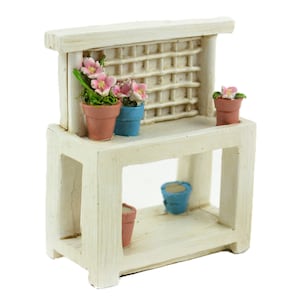 May include: A miniature white potting bench with a lattice back and a lower shelf. It features several small terracotta and blue pots, some holding pink flowers. The bench is designed for a fairy garden or similar decorative display.