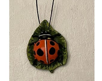 Può includere: Una coccinella rossa e nera con punti bianchi è posata su una foglia verde. La coccinella ha antenne nere ed è fatta di ceramica.