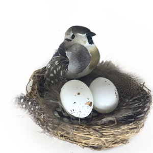 May include: A brown bird figurine sitting in a nest with two white speckled eggs. The nest is made of twigs and feathers.