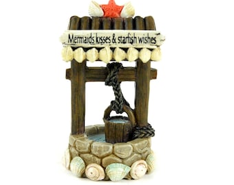 May include: A miniature wishing well figurine with a rustic wooden frame, stone base, and a bucket hanging from a rope. The roof is adorned with seashells and a red starfish. The sign reads "Mermaids kisses & starfish wishes".