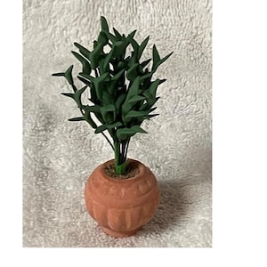 May include: A miniature artificial plant in a terracotta-coloured pot. The plant has dark green, pointed leaves and is set in a round, textured pot. The pot features a detailed, carved design around its base.