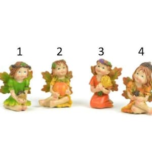 May include: Four small fairy figurines with wings, each holding a different autumnal item. The fairies are wearing green, yellow, orange, and brown dresses and have brown hair. They are sitting on a white surface.