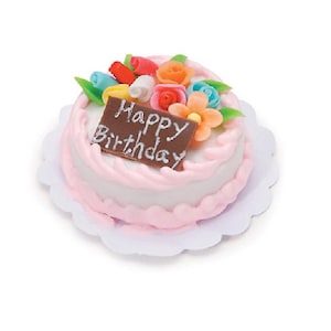 Puede incluir: Un pequeño pastel redondo con glaseado blanco y bordes rosados, decorado con flores miniatura de colores y una placa rectangular de chocolate marrón con el texto blanco Happy Birthday, sobre un tapete de papel blanco festoneado.