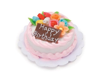 May include: A small round cake with white frosting and pink piped borders, topped with colorful miniature flowers and a rectangular brown chocolate plaque featuring the white text Happy Birthday, all resting on a white scalloped paper doily.