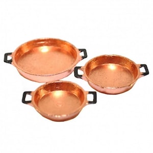 May include: Set of three copper-colored frying pans with black handles. The pans are different sizes and are arranged in a row from largest to smallest.