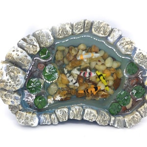 May include: A miniature stone pond with a blue resin water surface, featuring several colorful koi fish and green lily pads. The pond is surrounded by gray rocks.