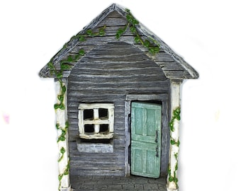 May include: A small, gray wooden house with a green door and a window. The house has a white trim and green vines growing up the sides.