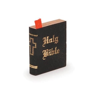 May include: A miniature black book with gold lettering that reads "Holy Bible." A gold cross is on the spine. A red bookmark is inserted. The book has a brown top edge.
