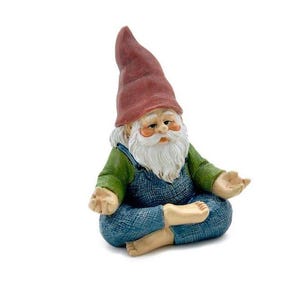May include: A small, red-hatted gnome figurine sits in a meditative pose with its legs crossed and hands resting on its knees. The gnome is wearing a green shirt and blue overalls.