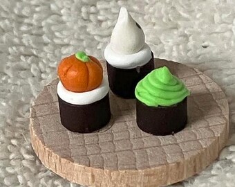 Könnte beinhalten: Miniatur-Halloween-Cupcakes auf einer kleinen Holzplattform. Die Cupcakes zeigen einen Kürbis, einen Geist und grünes Frosting. Die Cupcakes stehen auf einer runden, hellbraunen Plattform mit strukturierter Oberfläche.