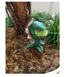 May include: A small green and pink plant figurine with a toothy mouth and one pink eye. The figurine is in a brown pot and is sitting on a bed of brown moss.
