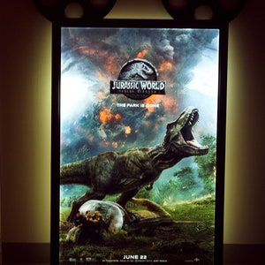 May include: A movie poster for Jurassic World: Fallen Kingdom, featuring a large, green Tyrannosaurus Rex dinosaur with its mouth open, roaring. The poster has a dark, stormy background with a fiery orange glow. The text "Jurassic World: Fallen Kingdom" is in white letters with a black outline, and the text "The Park is Gone" is in white letters below the title. The poster also includes the release date "June 22" in white letters.