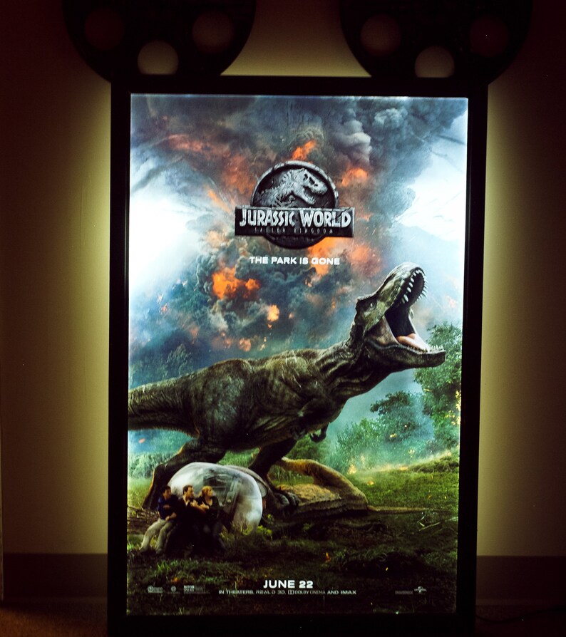 May include: A movie poster for Jurassic World: Fallen Kingdom, featuring a large, green Tyrannosaurus Rex dinosaur with its mouth open, roaring. The poster has a dark, stormy background with a fiery orange glow. The text "Jurassic World: Fallen Kingdom" is in white letters with a black outline, and the text "The Park is Gone" is in white letters below the title. The poster also includes the release date "June 22" in white letters.