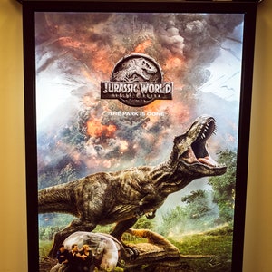 May include: A movie poster for Jurassic World: Fallen Kingdom. The poster features a large, roaring Tyrannosaurus Rex in front of a fiery, cloudy background. The text "Jurassic World: Fallen Kingdom" and "The Park is Gone" are visible on the poster.