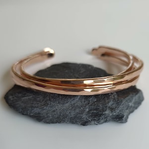 May include: Two rose gold hammered metal bangles stacked on a gray stone. The bangles are simple and elegant.