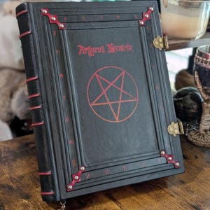 Könnte beinhalten: Ein schwarzes, ledergebundenes Tagebuch mit roten Akzenten und einem Pentagramm-Design. Das Tagebuch trägt den Titel "Argryros Necatrix".