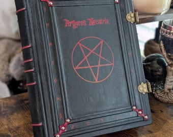 Peut inclure: Un journal relié en cuir noir avec des accents rouges et un motif de pentagramme. Le journal est intitulé "Argryros Necatrix".