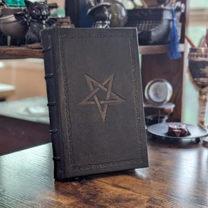 May include: A dark brown leather-bound book with a pentagram design embossed on the cover. The book has decorative border details and a ribbon bookmark. The book is displayed on a wooden surface.