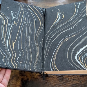 May include: Open journal with black covers and marbled pages. The pages feature a swirling pattern of black, white, and gold. The journal is open, revealing the inside pages, and has a black ribbon bookmark.