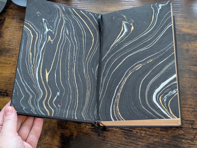 May include: Open journal with black covers and marbled pages. The pages feature a swirling pattern of black, white, and gold. The journal is open, revealing the inside pages, and has a black ribbon bookmark.