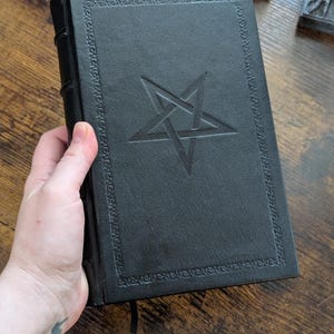 May include: A black book with a pentagram design embossed on the cover. The book has a decorative border and a black ribbon bookmark. The book is held in a hand.