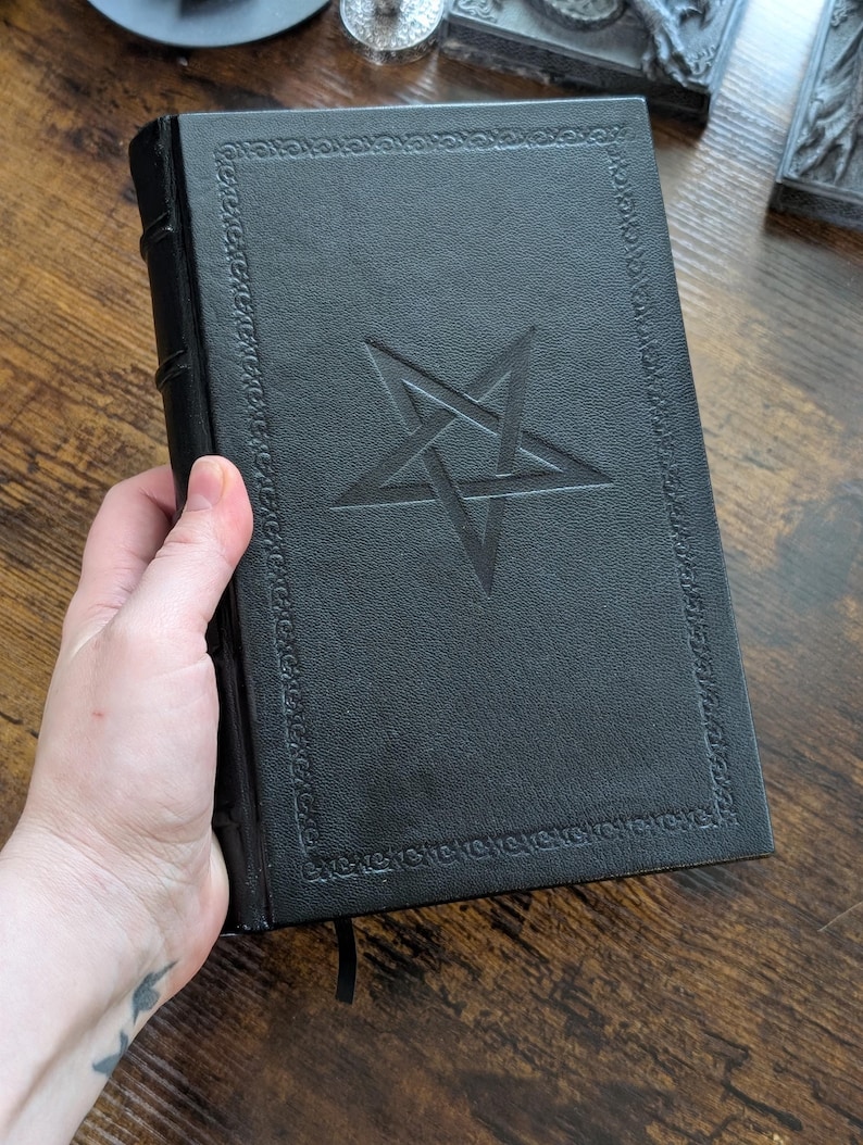 May include: A black book with a pentagram design embossed on the cover. The book has a decorative border and a black ribbon bookmark. The book is held in a hand.
