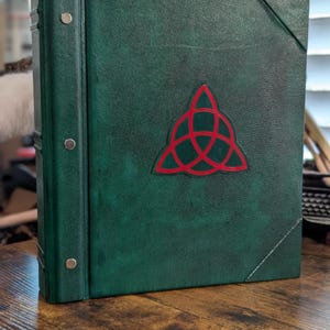 May include: A green leather-bound book with a red Celtic knot emblem. The book has silver metal accents on the spine and a diagonal stitched corner. The book is sitting on a wooden surface.