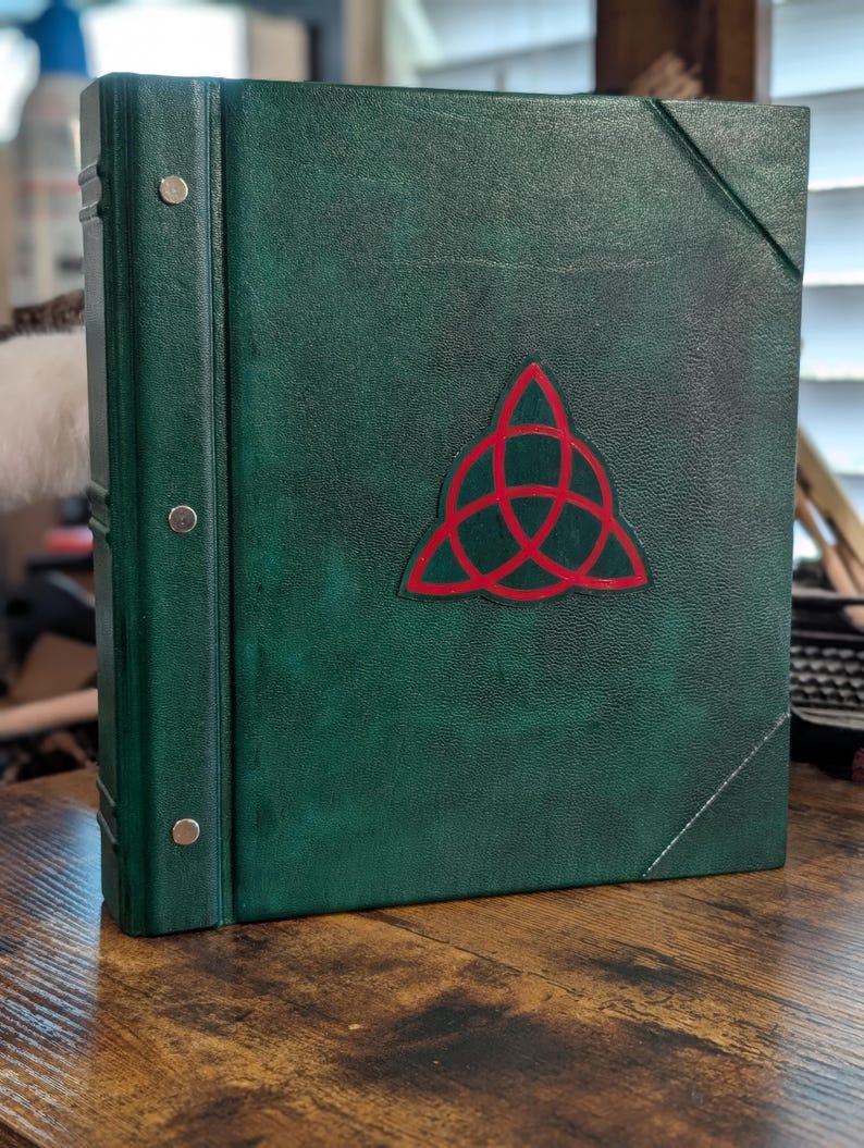 May include: A green leather-bound book with a red Celtic knot emblem. The book has silver metal accents on the spine and a diagonal stitched corner. The book is sitting on a wooden surface.