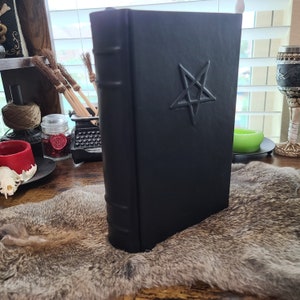 May include: A black book with a pentagram embossed on the cover. The book has a textured appearance and is standing upright. The spine has raised bands. The book is on a fur-like surface.