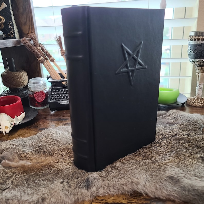 May include: A black book with a pentagram embossed on the cover. The book has a textured appearance and is standing upright. The spine has raised bands. The book is on a fur-like surface.