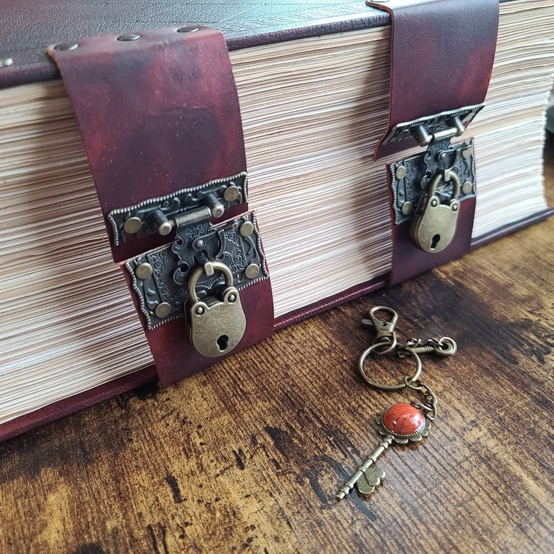 K&ouml;nnte beinhalten: Ein dickes, ledergebundenes Buch mit zwei verzierten Metallschl&ouml;ssern und einem Schl&uuml;ssel mit einem roten Steinanh&auml;nger. Das Buch ist geschlossen und hat einen rustikalen, Vintage-Look.