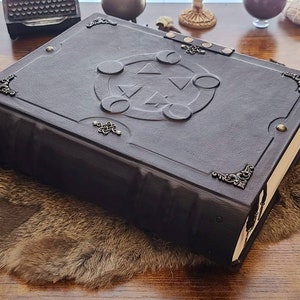 May include: A dark brown leather-bound journal with a decorative silver clasp and a silver embossed design on the cover. The design features a circle with a triangle inside and three crescent moons around it.