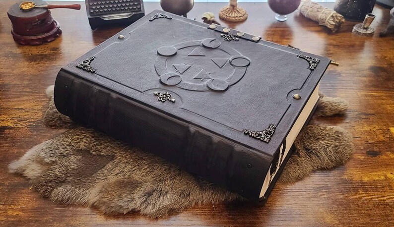 May include: A dark brown leather-bound journal with a decorative silver clasp and a silver embossed design on the cover. The design features a circle with a triangle inside and three crescent moons around it.