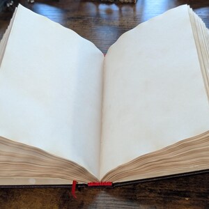 Puede incluir: Un libro abierto con p&aacute;ginas en blanco de color blanquecino. Tiene una cubierta oscura y un marcador de cinta roja. Las p&aacute;ginas parecen envejecidas, d&aacute;ndole un aspecto vintage. El libro est&aacute; sobre una superficie de madera.