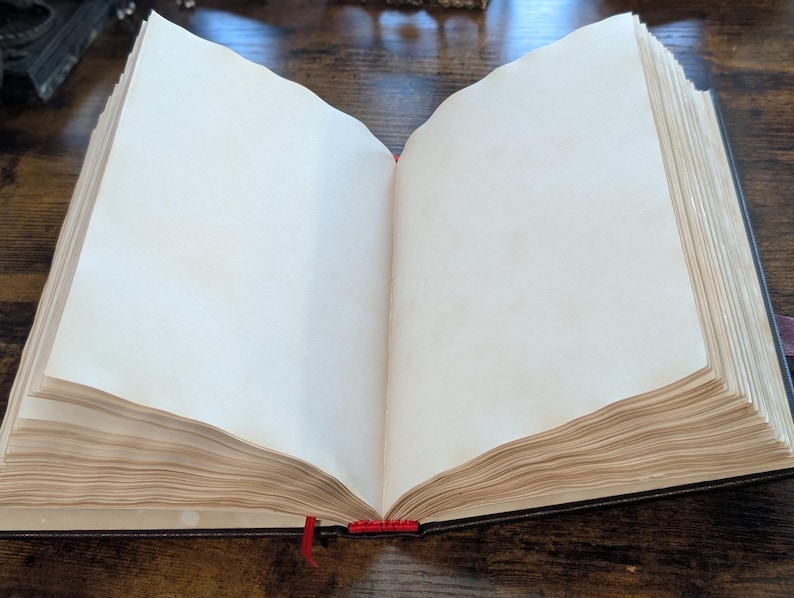 Puede incluir: Un libro abierto con p&aacute;ginas en blanco de color blanquecino. Tiene una cubierta oscura y un marcador de cinta roja. Las p&aacute;ginas parecen envejecidas, d&aacute;ndole un aspecto vintage. El libro est&aacute; sobre una superficie de madera.