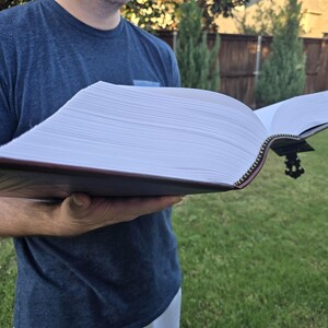 May include: A large, leather-bound book with a zipper closure is open to reveal hundreds of blank pages. The book is held by a person in a blue shirt.