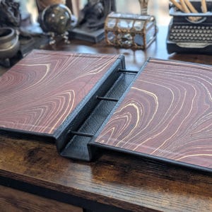 May include: An open, vintage-style journal with a marbled, reddish-brown cover and black binding. The cover features a swirling pattern of light beige and brown, giving it a classic, antique look. The journal is displayed open on a wooden surface.