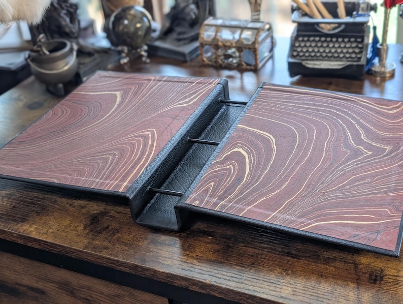 May include: An open, vintage-style journal with a marbled, reddish-brown cover and black binding. The cover features a swirling pattern of light beige and brown, giving it a classic, antique look. The journal is displayed open on a wooden surface.