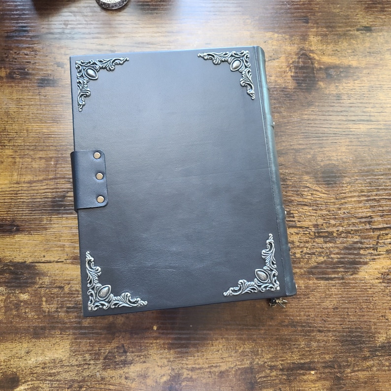 May include: A black leather journal with decorative silver corner embellishments and a leather strap closure. The journal is closed and rests on a wooden surface. The corners feature a vintage design.