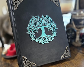 May include: Black leather journal with a teal tree of life design embossed on the front cover. The journal has decorative gold corners.