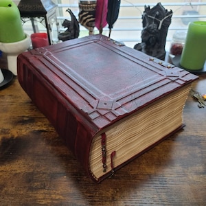 K&ouml;nnte beinhalten: Ein gro&szlig;es, rot gebundenes Buch mit dekorativen Metallakzenten und einem abgenutzten, alten Aussehen. Das Buch ist geschlossen und hat einen dicken R&uuml;cken mit vielen Seiten.