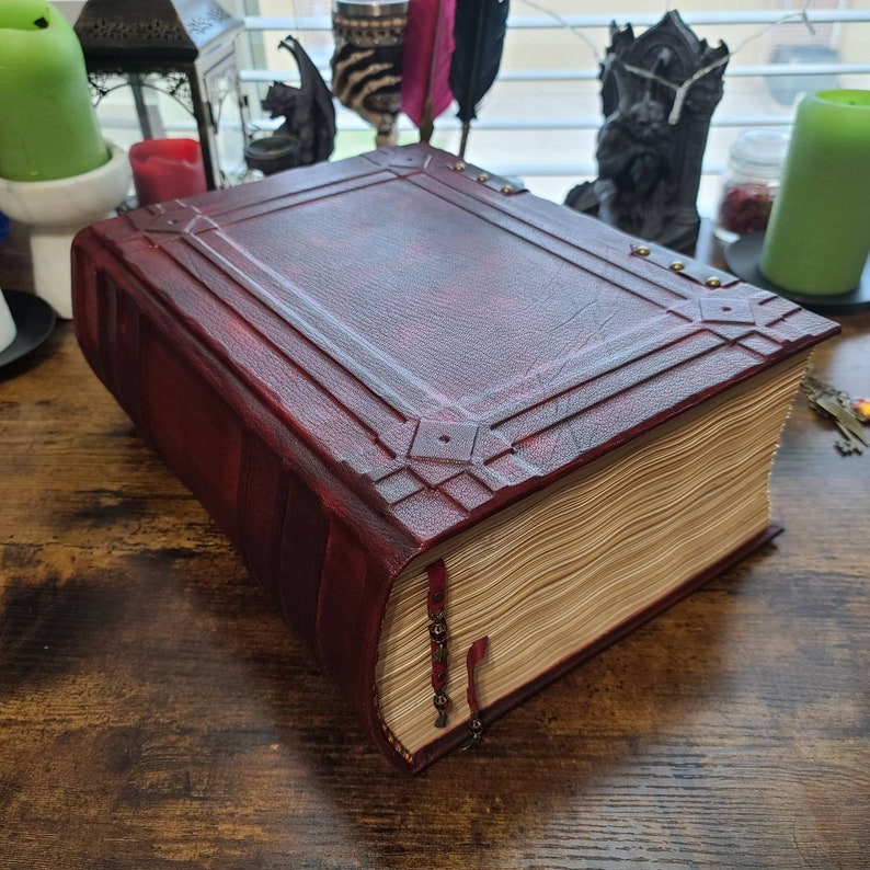 K&ouml;nnte beinhalten: Ein gro&szlig;es, rot gebundenes Buch mit dekorativen Metallakzenten und einem abgenutzten, alten Aussehen. Das Buch ist geschlossen und hat einen dicken R&uuml;cken mit vielen Seiten.