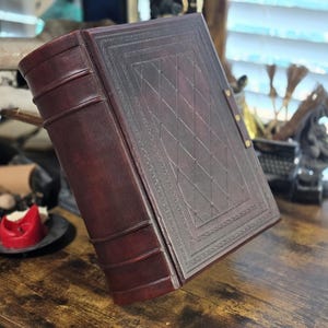 May include: A dark brown leather-bound journal with a decorative embossed pattern on the cover. The journal has a spine with raised bands and is closed.