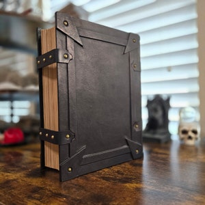 May include: A black leather-bound book with brass accents and a worn, aged appearance. The book is closed and standing upright on a wooden surface.