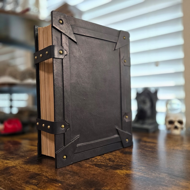May include: A black leather-bound book with brass accents and a worn, aged appearance. The book is closed and standing upright on a wooden surface.